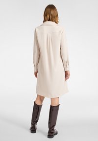 Beige Langarmkleid mit Kragen, ausgestattet mit einem Falten-Detail im Rücken und kombiniert mit glänzenden knielangen schwarzen Stiefeln. Glattes Material.