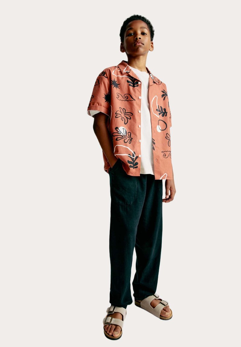 Camicia a maniche corte in terracotta con motivi floreali neri, abbinata a pantaloni scuri larghi e sandali chiari. Design semplice con bottoni.