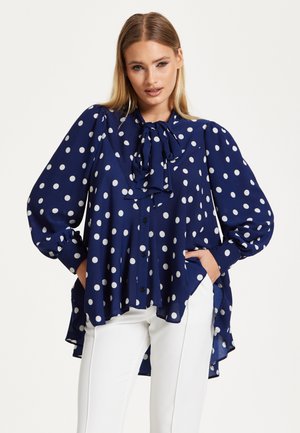 Navy blue blouse with white polka dots, long puff sleeves, tie neck detail, and asymmetric hem. Fabric is lightweight and flowing.
