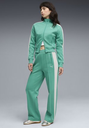 Γυναίκα με tracksuit Puma σε μέντα πράσινο χρώμα με λευκές ρίγες και ασημένια αθλητικά παπούτσια, στέκεται μπροστά σε γκρίζο φόντο με βαθμίδωση.