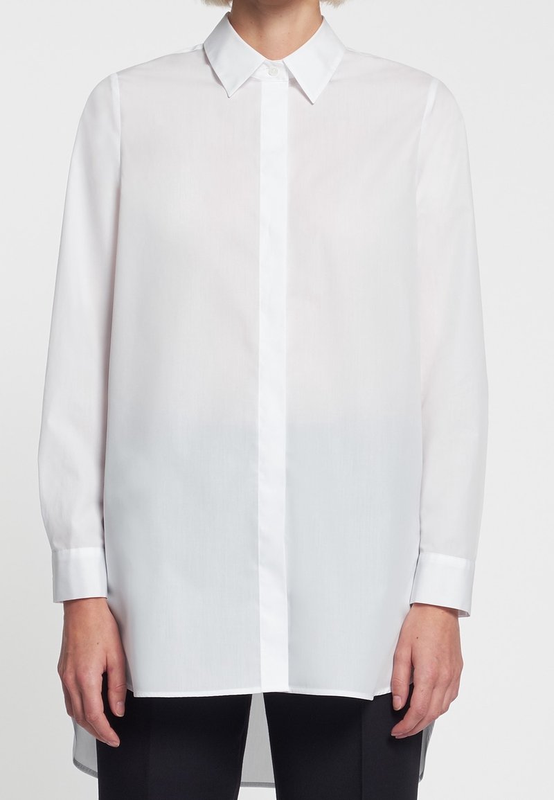 Personne portant une chemise blanche boutonnée à manches longues avec un col pointu et un pantalon noir, visible du cou aux hanches.