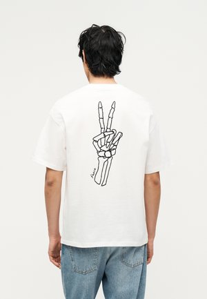 Persona che indossa una maglietta bianca con una mano scheletrica nera che fa il segno di pace sulla schiena, abbinata a jeans blu.