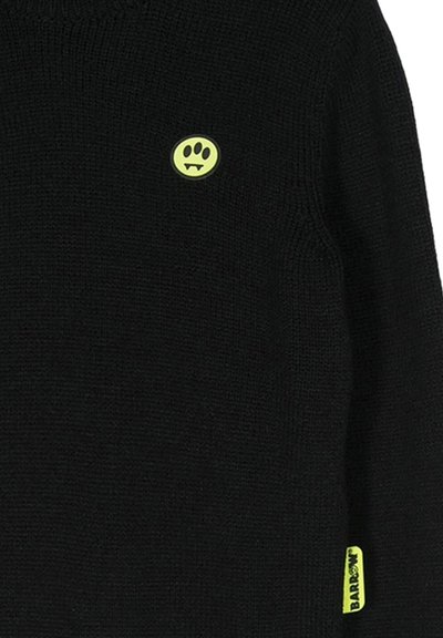 Maglione nero a maglia con un piccolo logo circolare giallo con tre occhi e zanne, e un'etichetta gialla sull'orlo.