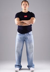 Dunkelblaues T-Shirt mit rotem Etikett, hellblaue Jeans und weiße Sneakers. Das Model steht mit verschränkten Armen und präsentiert einen lässigen Streetwear-Stil.