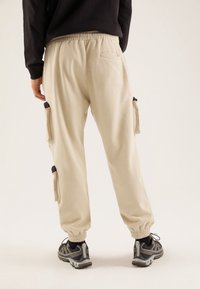 Person, der beigefarbene Cargo-Jogginghose und schwarze Sneakers trägt, steht mit dem Rücken zu einem einfarbigen hellen Hintergrund.