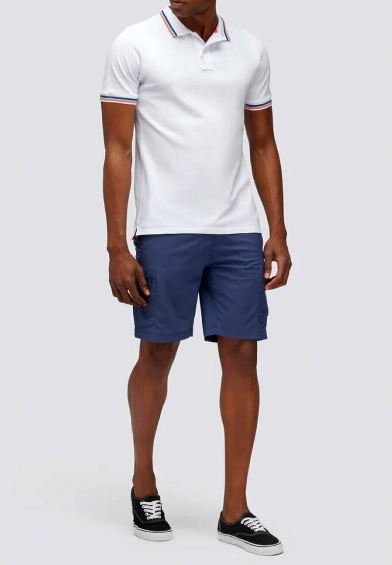 Polo bianco con colletto colorato; abbinato a pantaloni corti blu navy. Sneakers nere con lacci bianchi. Design semplice e tessuto leggero.