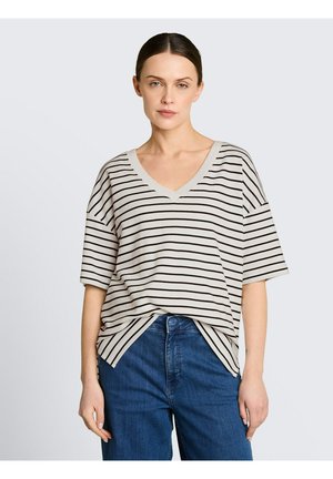 Kobieta o ciemnych włosach, związanych w niski kok, ubraną w biały T-shirt z czarnymi poziomymi paskami i niebieskie dżinsy, stojąca na jednolitym tle.