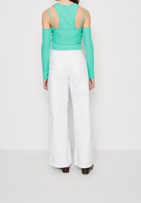 Nainen, joka pitää yllään turkoosia kyljistä avoimia ribbipaitaa ja korkeavyötäröisiä valkoisia leveitä housuja, seisoo selkä kameran suuntaan.