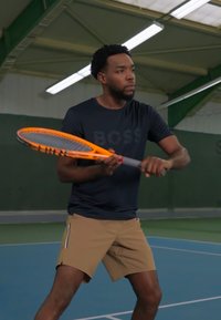 Teniszjátékos narancssárga csíkos ütőt tart, fekete "BOSS" logós pólót és bézs sortot visel, egy kék pályán áll.