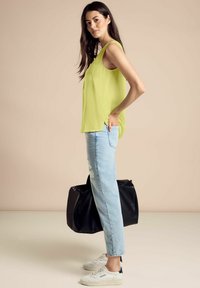 Blusa senza maniche verde lime con spacco laterale, abbinata a jeans chiari strappati e sneakers bianche, mentre tiene una borsa nera.