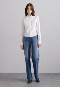 Cămașă albă cu nasturi, având două buzunare în față, asortată cu blugi denim drepti de culoare albastră deschis. Poartă pantofi cu toc negri.
