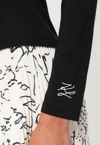 Must valge tikandiga pikade varrukatega pluus; kantud valge laskpliseeritud seeliku kohal, millel on mustad abstraktsed mustrid.