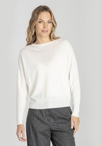 Blondynka w luźnej białej bluzie z długim rękawem i szarych spodniach z wysokim stanem, stojąca na tle jasnego, jednolitego tła.