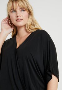 Svart, løst tilpasset topp med V-hals, korte vide ermer, myk tekstur og drapert design. Har en enkelt knapp ved halsen.