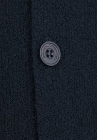 Ciemnoniebieska dzianinowa tkanina swetra z teksturową powierzchnią oraz czarnym guzikiem z czterema otworami z wytłoczonym napisem "PAKOLORIENTE."