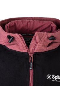 Giacca esterna nera con cappuccio foderato in fleece rosa. Presenta una chiusura frontale con zip, un tirante e un logo sul lato inferiore destro.