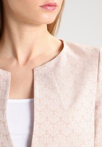 Giacca rosa chiaro con un motivo testurizzato a forme di ventaglio, caratterizzata da un collo rotondo e un design aperto nella parte frontale. Il materiale appare liscio.
