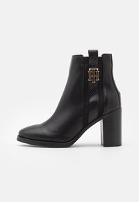 Bottine en cuir noir avec un bout pointu, un panneau latéral texturé et un accent logo doré. Présente un talon moyen bloc et une finition lisse.