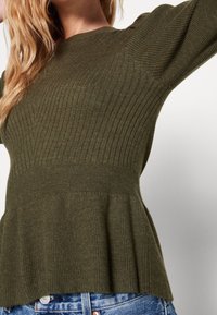 Maglione a coste verde oliva con corpetto aderente e orlo a peplum. Caratterizzato da maniche lunghe e dettagli testurizzati. Indossato con jeans di denim.