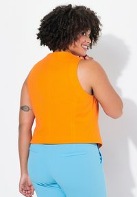 Oranje mouwloze top met een aansluitend ontwerp, met een gladde textuur. Gecombineerd met licht blauwe broek, waarbij details van zijzakken zichtbaar zijn.