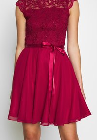Robe rouge sans manches avec corsage en dentelle, paillettes, et ceinture en ruban de satin nouée en un nœud à la taille sur une jupe fluide en chiffon.