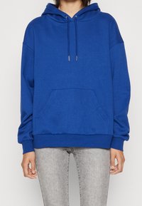 Felpa blu in pile con tasca frontale, cappuccio con cordino e polsini a coste. Indossata con jeans grigi, che hanno una vestibilità ampia e rilassata.