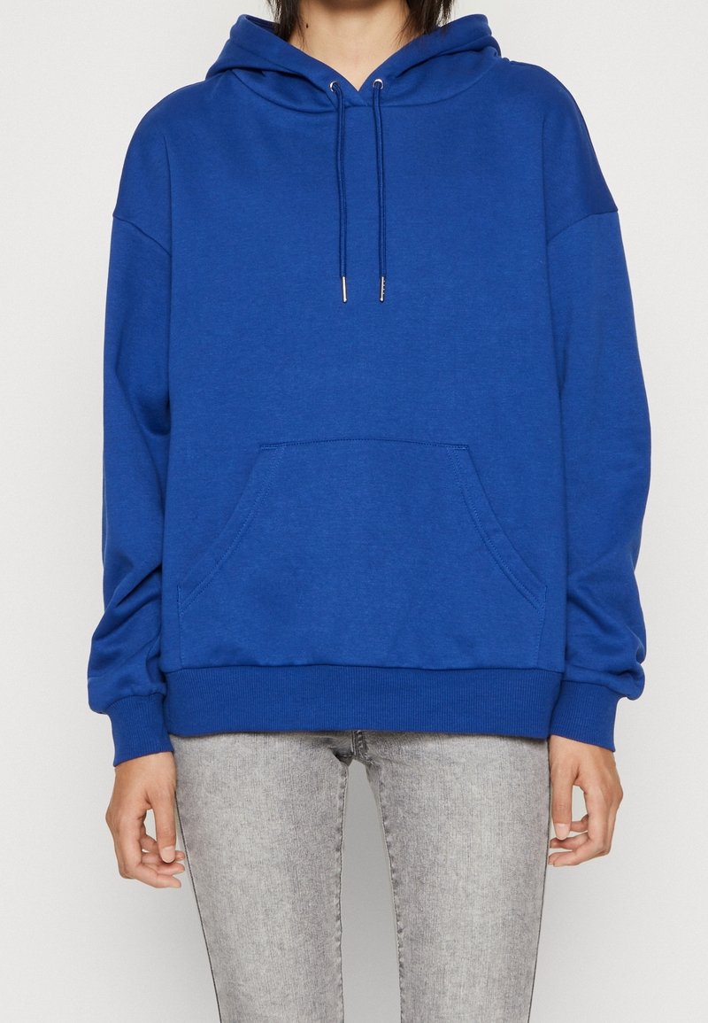 Felpa blu in pile con tasca frontale, cappuccio con cordino e polsini a coste. Indossata con jeans grigi, che hanno una vestibilità ampia e rilassata.