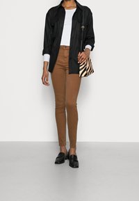 Camisa negra abotonada, blusa blanca, pantalones ajustados marrones y mocasines negros. Un bolso de estampado de cebra acentúa el atuendo. Texturas suaves a lo largo.