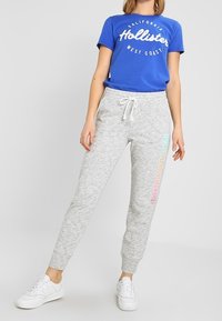 Grå joggingsbyxor med elastisk midja och dragsko, med "Hollister"-logotyp i pastellfärger. Matchas med en blå grafisk t-shirt.
