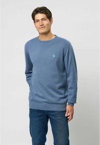 Lyseblå sweater med ribbet rund hals, tekstureret stof og lille logo-detalje på brystet, parret med blå jeans.