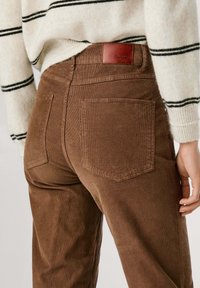 Bruine corduroy broek met een subtiele ribbelstructuur, met twee achterzakken en een leren label op de tailleband. Gestreepte trui zichtbaar erboven.