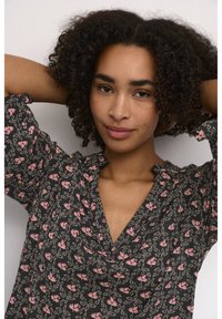 Blouse noire avec un motif floral rose, présente une texture douce, un décolleté en V, des manches courtes et des fronces aux coutures des épaules.