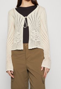 Krämfärgad stickad cardigan med mönster av spets, V-ringning, knytanordning framtill och utsvängda ärmar. Kombinerad med bruna cargo-byxor.