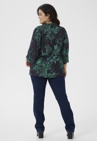 Blouse florale en vert foncé et noir, avec des manches 3/4 et une coupe décontractée. Associée à un jean bleu foncé et des bottes noires à talons.