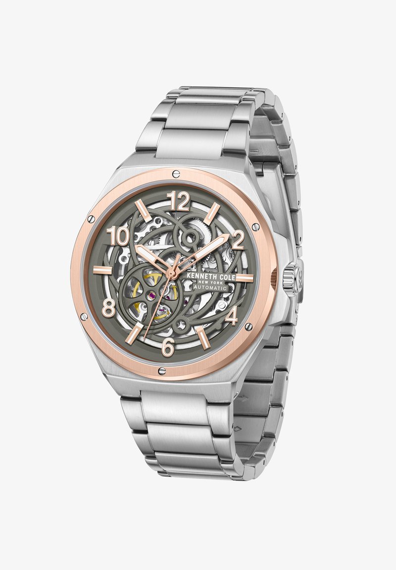 Reloj de acero inoxidable plateado con un bisel de oro rosa, esfera esqueleto y grandes números. Presenta intrincados engranajes y una pulsera de eslabones metálicos.