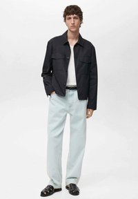 Zwarte, lichte, knoopjacks met twee borstzakken, gecombineerd met lichtblauwe jeans en zwarte sandalen, op een witte achtergrond.