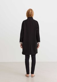 Juodas ilgas paltas su elegantiška siluetu ir aukšta apykakle, su vientisa tekstūra ir susuktais rankogaliais. Dėvimas ant įtemptų juodų kelnių ir sandalių.