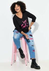 Černé tričko s výstřihem do V a růžovou grafikou "Jen se usmívej"; roztrhané modré džíny s barevnými nášivkami; černé kotníkové boty; sedící na růžovém stoličku.