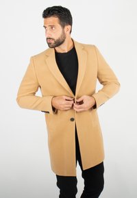 Manteau en laine beige avec un bouton unique, revers crantés et deux poches avant. Le modèle porte une chemise noire et ajuste la fermeture du manteau.