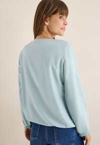 Himmelbluse mit langen Ärmeln und rundem Ausschnitt, glatter Textur und geradem Rücken. Kombiniert mit dunklen Jeans.