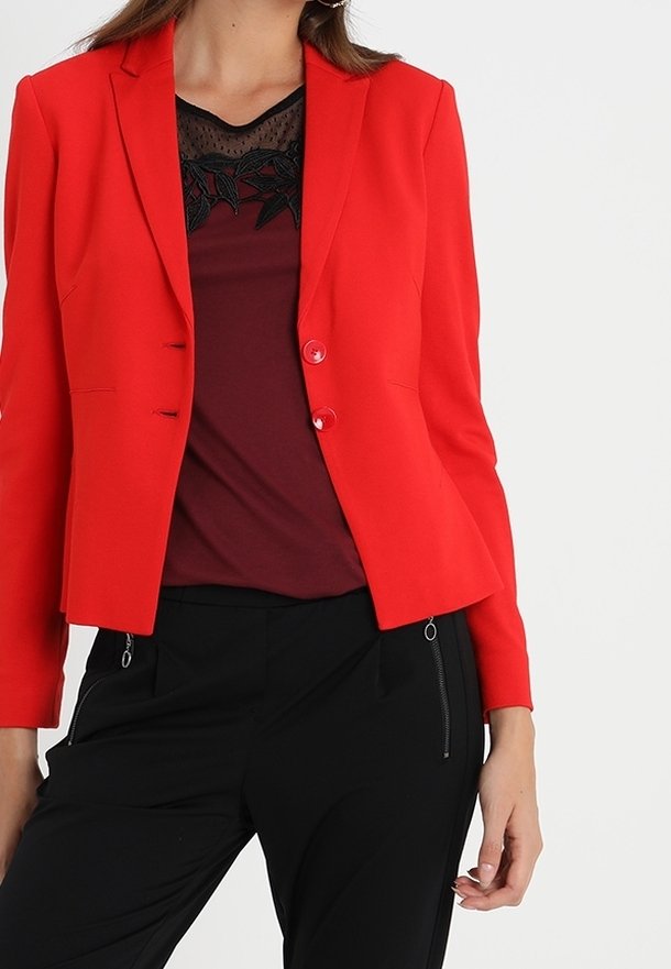 Veste rouge à manches longues, deux boutons et coupe ajustée. Portée sur un haut bordeaux avec des détails en dentelle. Associée à un pantalon noir.