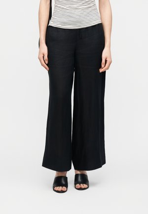 Femme portant un pantalon large noir, des sandales à talons ouvertes noires et un haut à manches courtes à rayures blanches et noires.