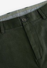 Mørkegrønne chinos lavet af et struktureret stof. Har en knaplukning, flad front og et mønstret indvendigt taljebånd.