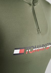 Grünes Polohemd mit einem Halb-Reißverschluss-Design, aus glattem Stoff gefertigt, mit einem schwarzen und roten "TOMMY SPORT"-Logo und kontrastierenden gestreiften Akzenten.