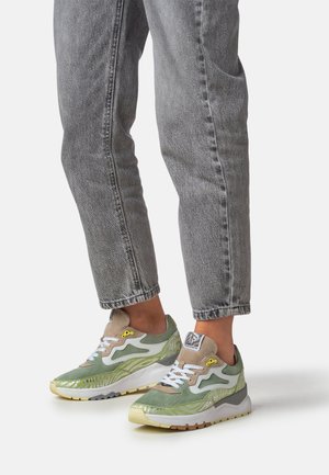 Person iført grå jeans og grønne, beige og hvide sneakers med hvide snørebånd, stående på en lys baggrund.