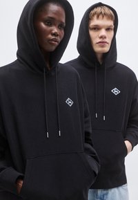 Čierne mikiny s kapucňou vyrobené z jemnej látky s nastaviteľnými šnúrkami, s malým diamantom v podobe loga na hrudi a predným vreckom.