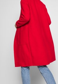 Persoon in een felrode wollen jas over lichtblauwe jeans, gezien van achteren tegen een effen grijze achtergrond.