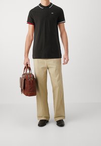 Czarna koszulka polo z białym kołnierzykiem i czerwonymi paskami przy mankietach, beżowe spodnie oraz brązowa skórzana torba z metalowymi detalami.