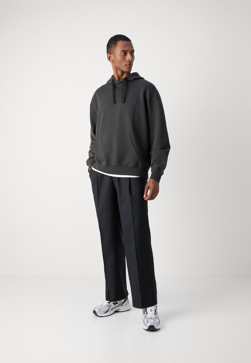 Donkergrijze hoodie met een zak aan de voorkant, gecombineerd met zwarte broeken en zilveren sneakers. De stof lijkt zacht en heeft een relaxed fit ontwerp.