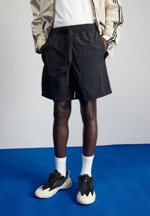 Person iført sorte shorts med bælte, hvide crew-sokker, sorte og hvide sneakers samt en let jakke med sorte striber, der står på blå gulv.